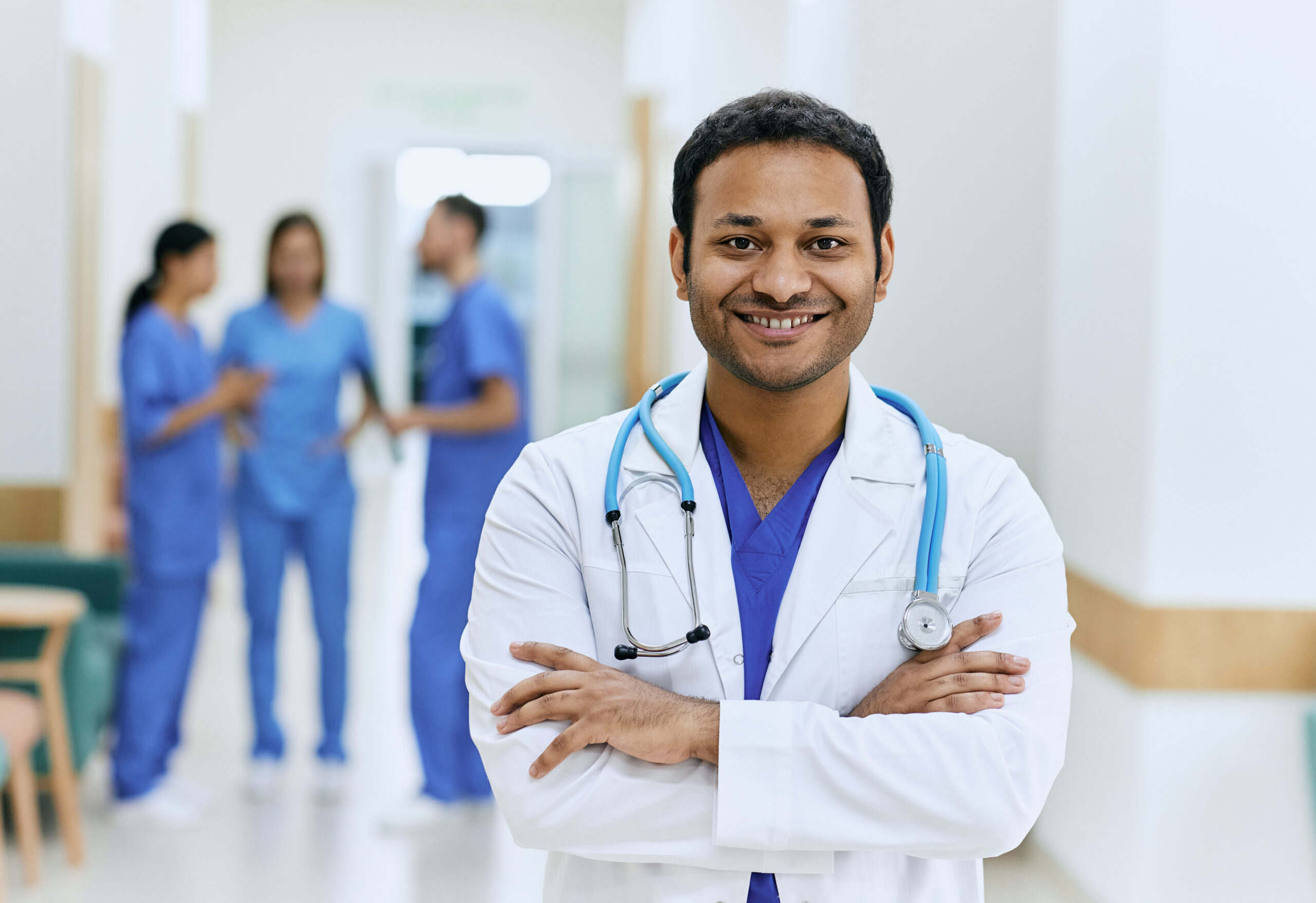 Portrait of experienced doctor from India with crossed arms and stethoscope standing in hallway of modern hospital. Medical profession